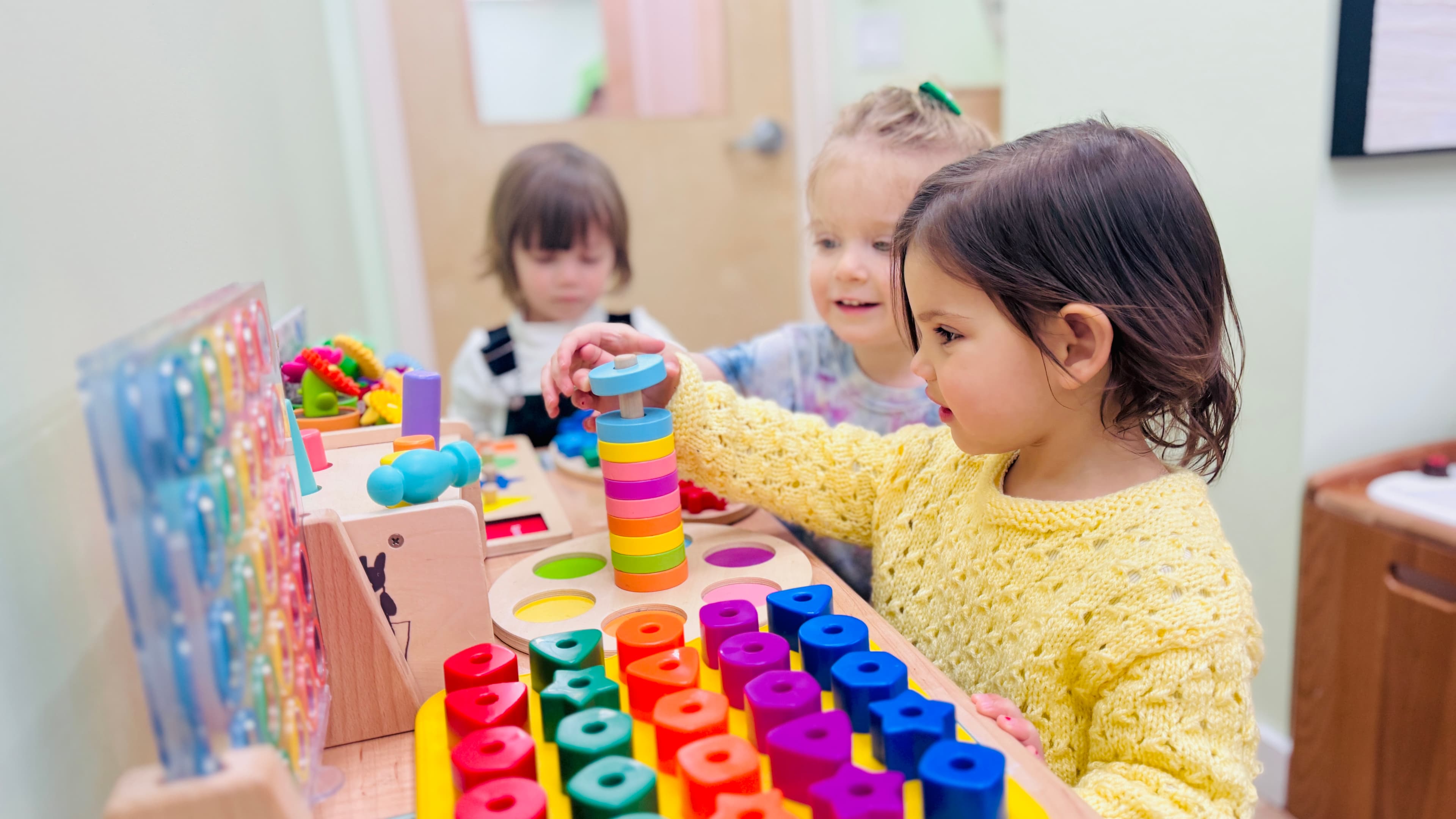 Three early preschoolers playing with colorful wooden Montessori sorting and stacking toys at a classroom table