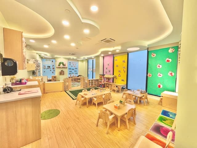 Bright toddler classroom with child-sized wooden tables and chairs, low Montessori shelving, and a colorful apple wall display