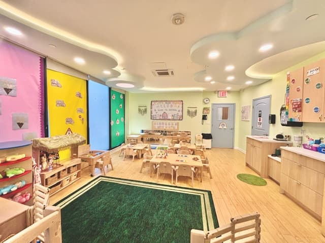 Toddler classroom with a green play rug, child-sized tables, wooden shelves, and a welcoming back-to-school display board