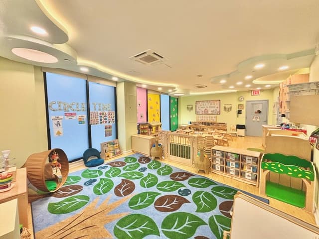 Cozy toddler classroom featuring a leaf-patterned rug, a Circle Time wall display, wooden cribs, and Montessori materials shelving