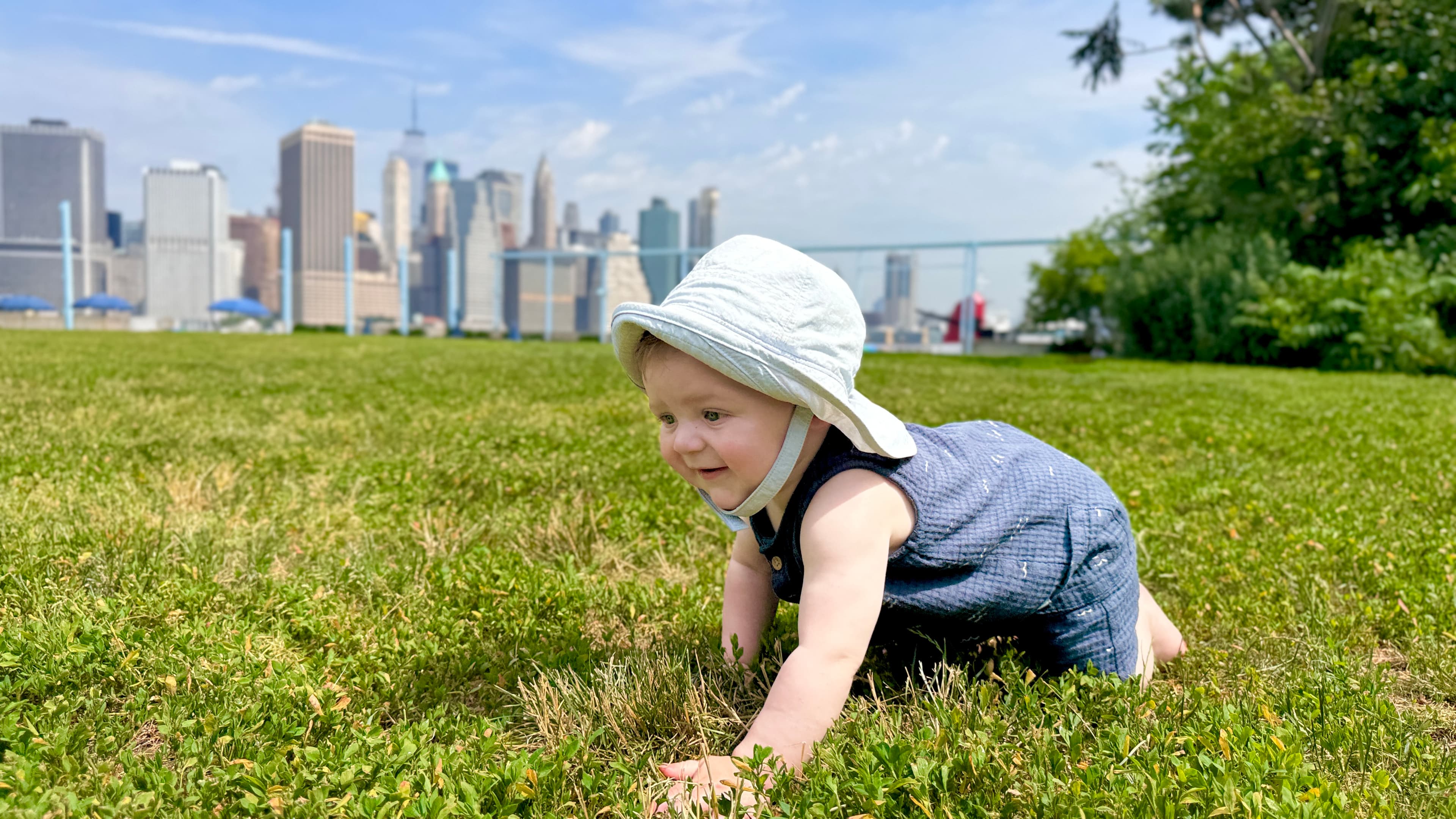 Infant in a sun hat crawling and exploring the grass at a Brooklyn park with the Manhattan skyline behind