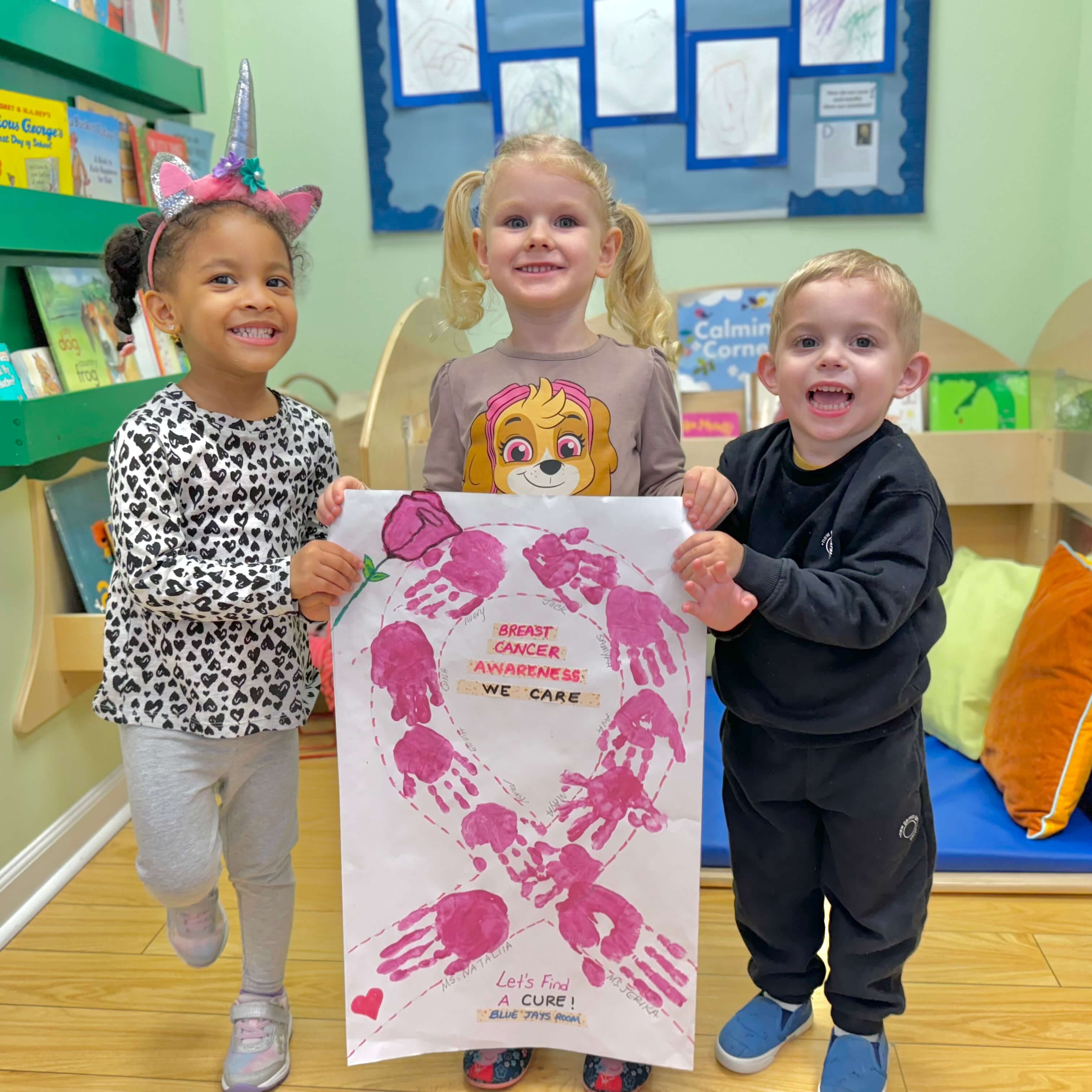 Pre-K children proudly holding a collaborative handprint art project in a book-filled classroom