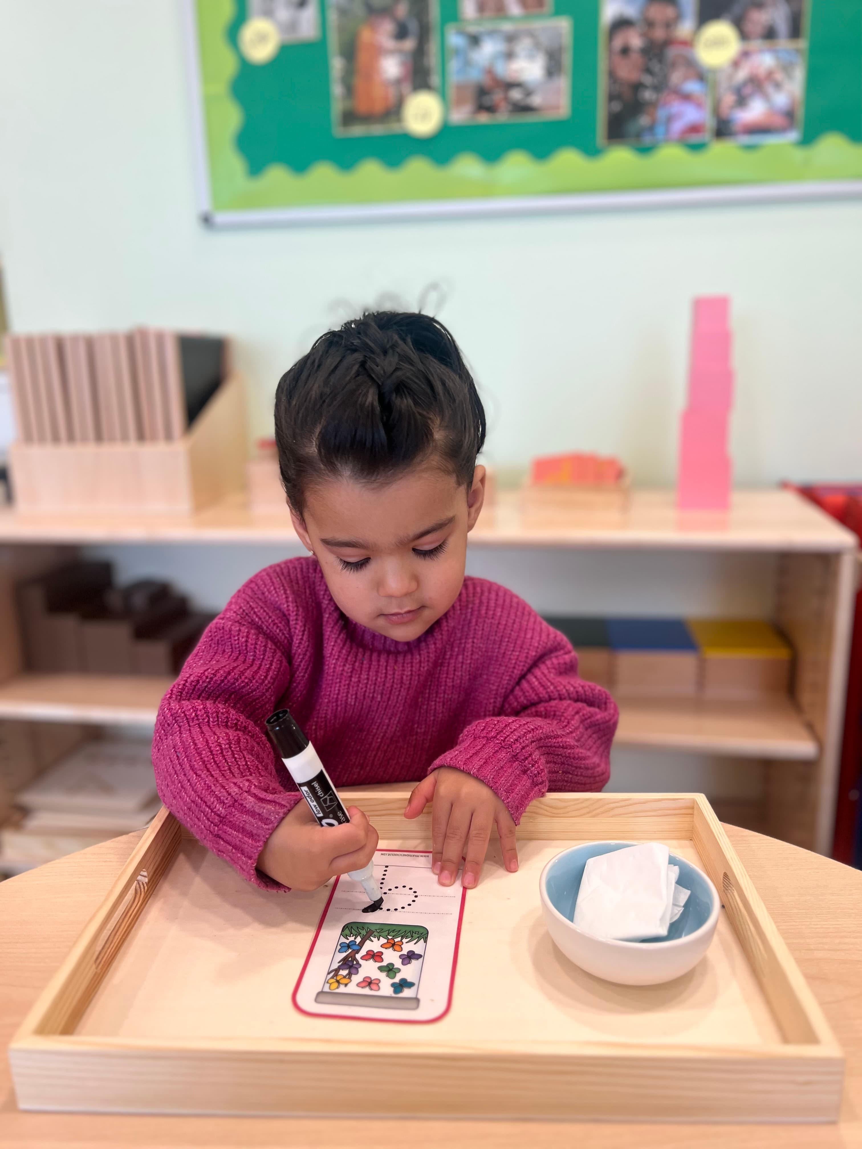 Pre-K child in a pink sweater tracing a number on a Montessori tracing worksheet at a wooden tray