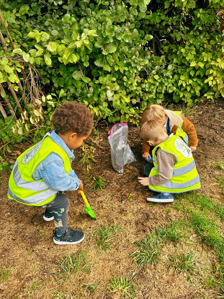 Pre-K children in safety vests exploring nature outdoors, digging and collecting specimens near the bushes