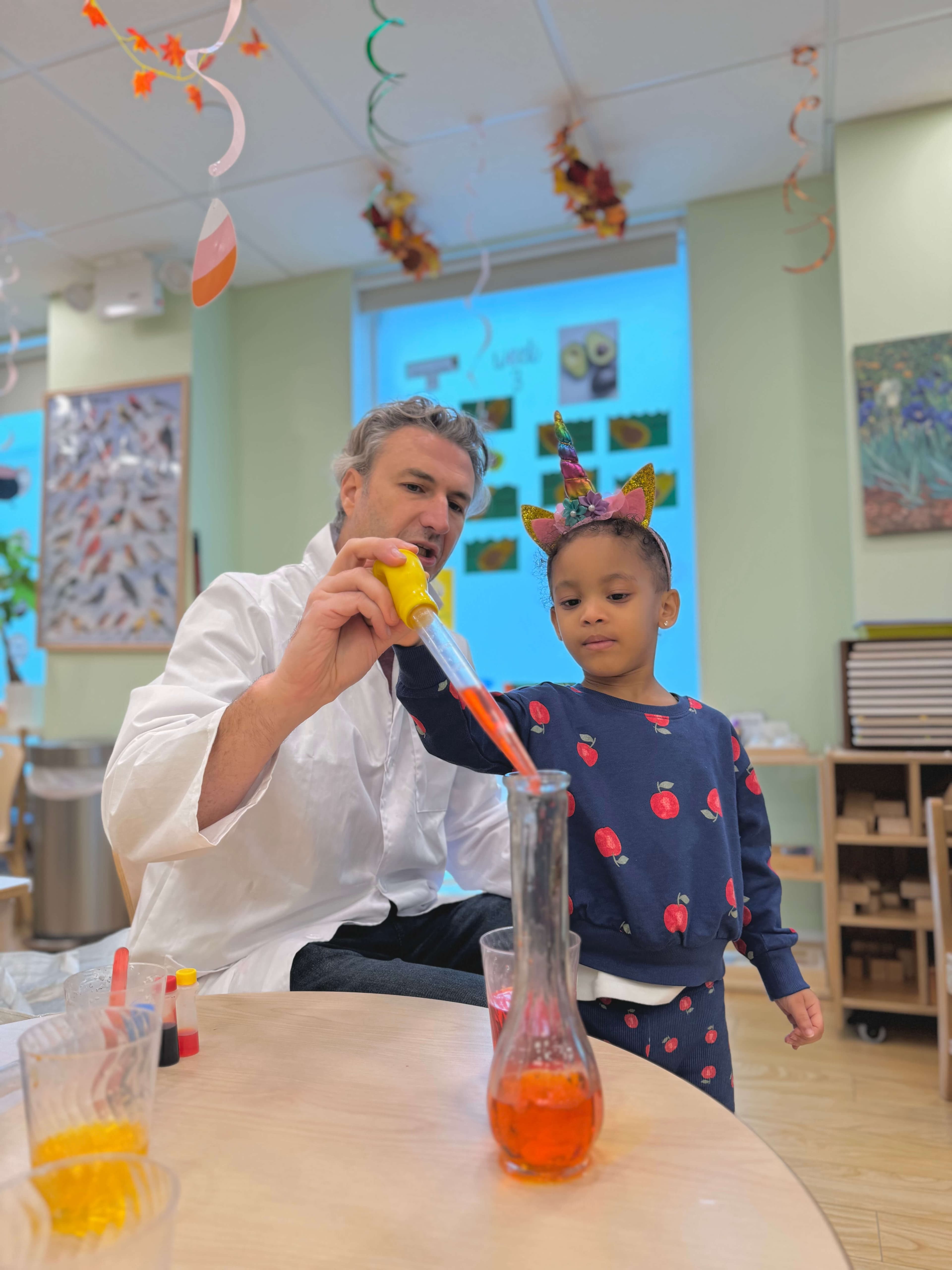 Pre-K child doing a hands-on science experiment with a teacher, using a pipette to add colored liquid to a flask