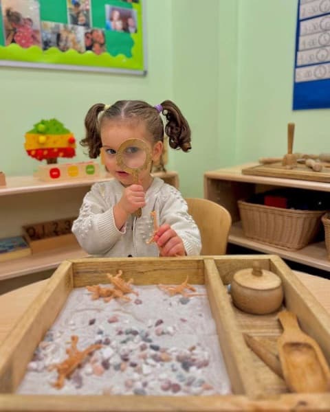Pre-K child using a magnifying glass to explore a Montessori sensory dig tray with sand, pebbles, and dinosaur figures