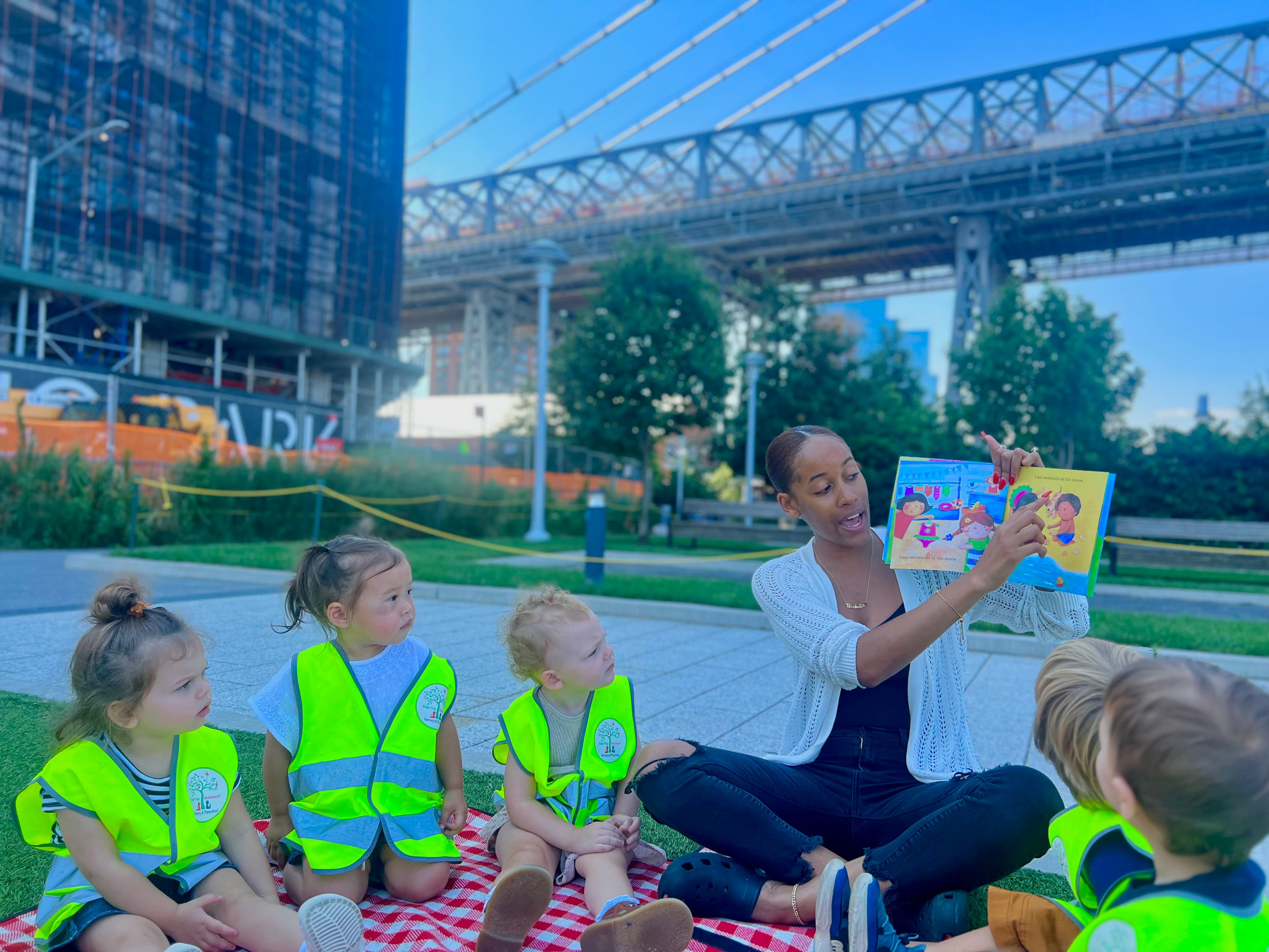 Teacher reading a picture book to toddlers in safety vests at the park under the Williamsburg Bridge