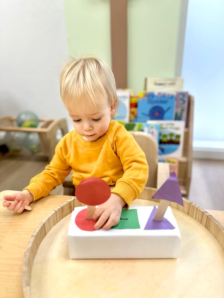 Toddler in a yellow sweater working on a Montessori shape-sorting activity at a wooden table