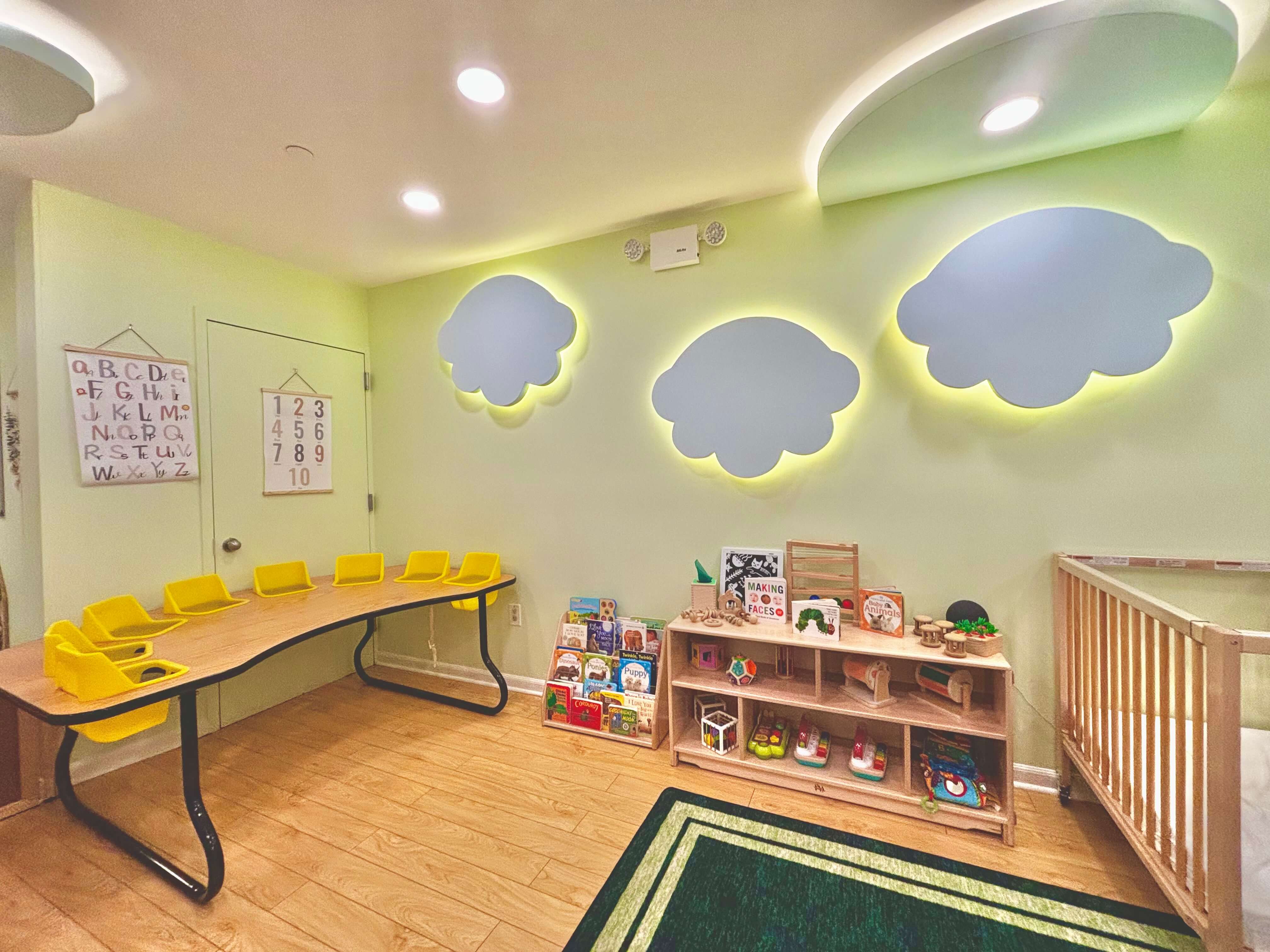 Bright infant classroom with cloud-shaped wall lights, Montessori book and materials shelf, and a wooden crib