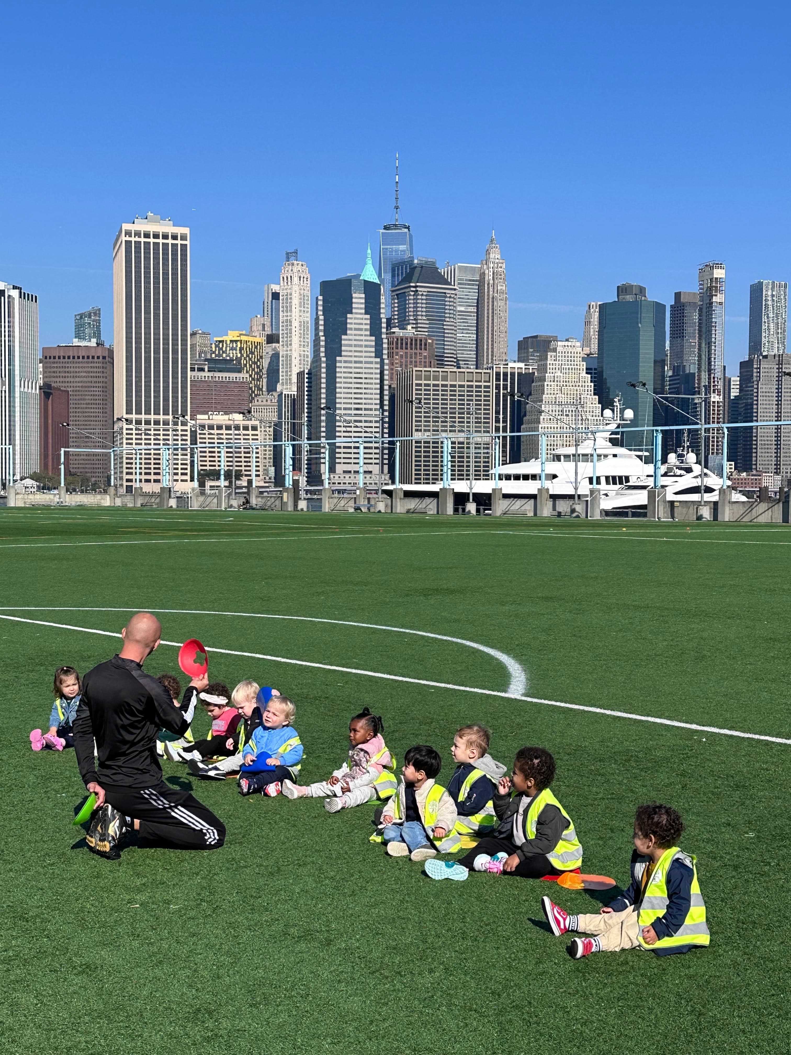 Coach leading a group of Kinder Prep campers in a field sports session with the Manhattan skyline behind them at Brooklyn Bridge Park