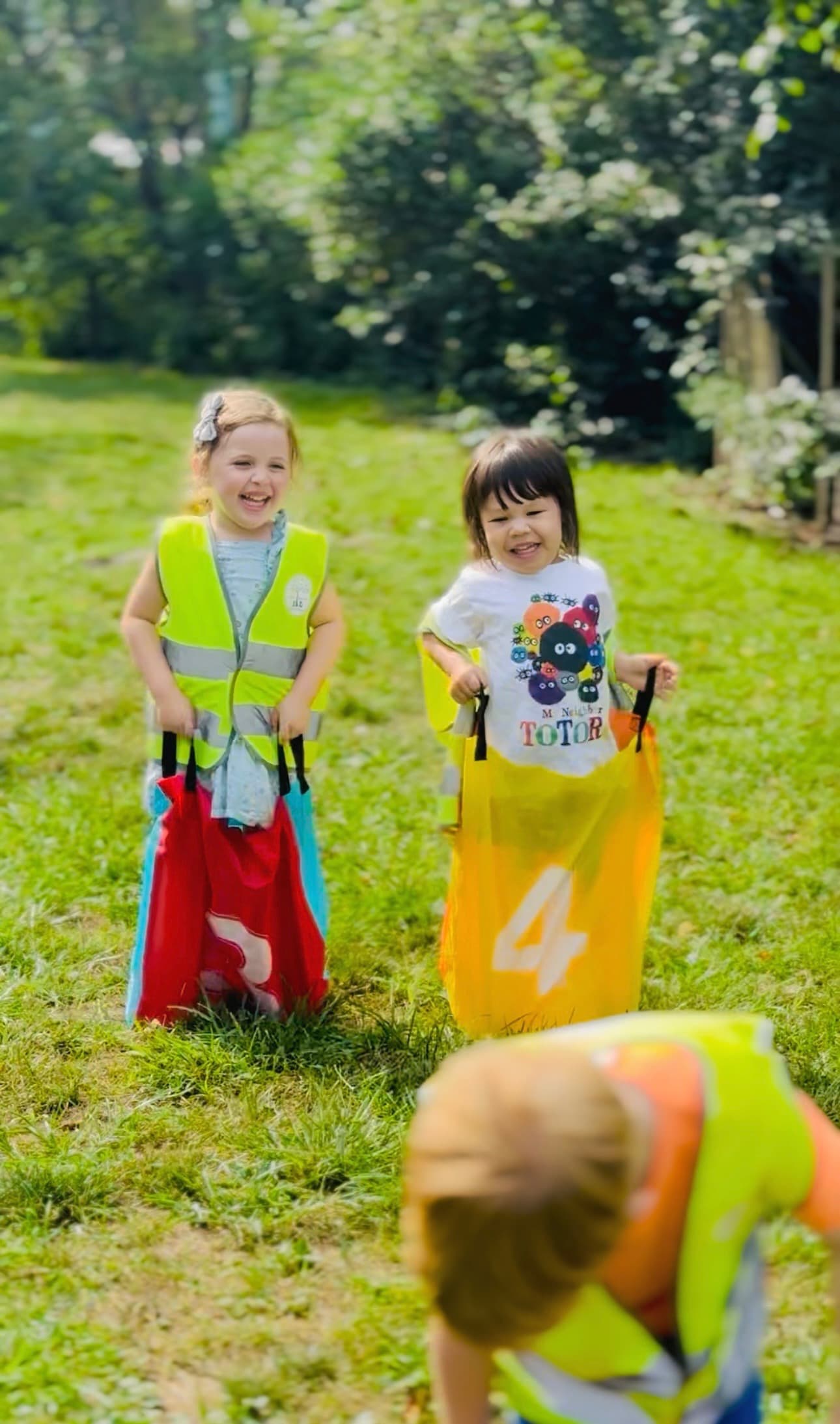 Two laughing Kinder Prep campers in safety vests racing in potato sacks on a sunny field at summer camp