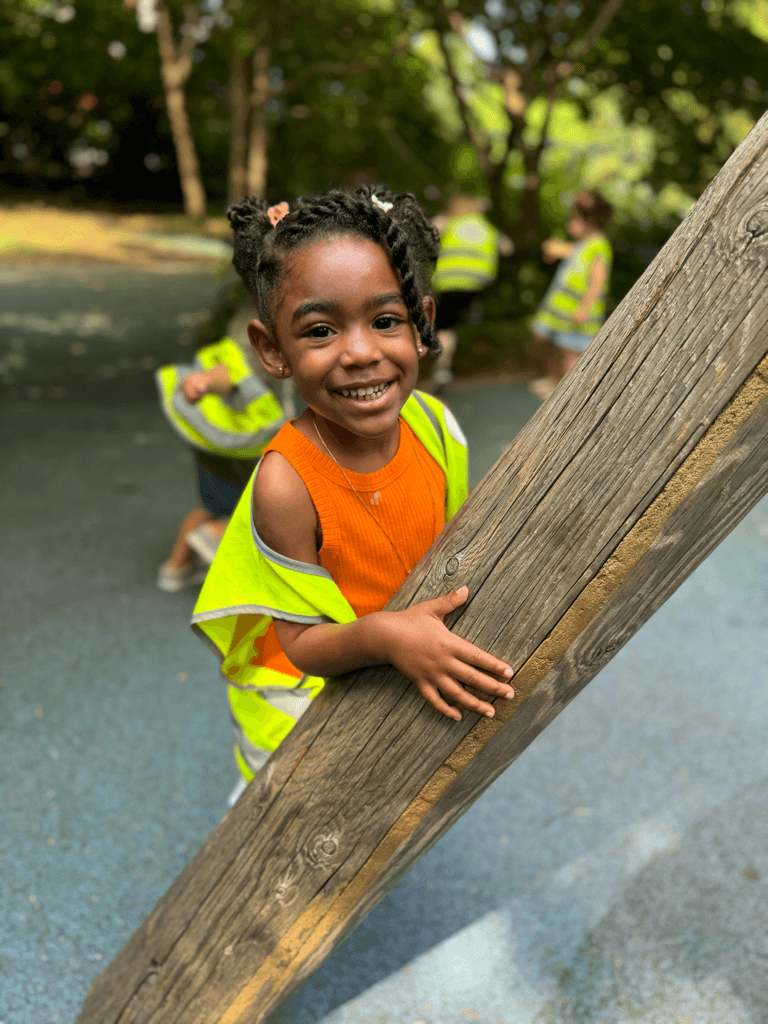 Smiling Kinder Prep camper hugging a wooden climbing log at the Pier 6 playground in Brooklyn Bridge Park
